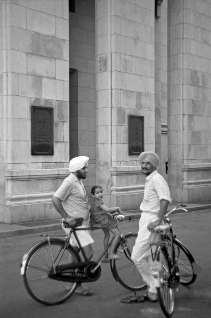 Two Asian men resting on their bicycles chatting in the street. Singapore, 1962