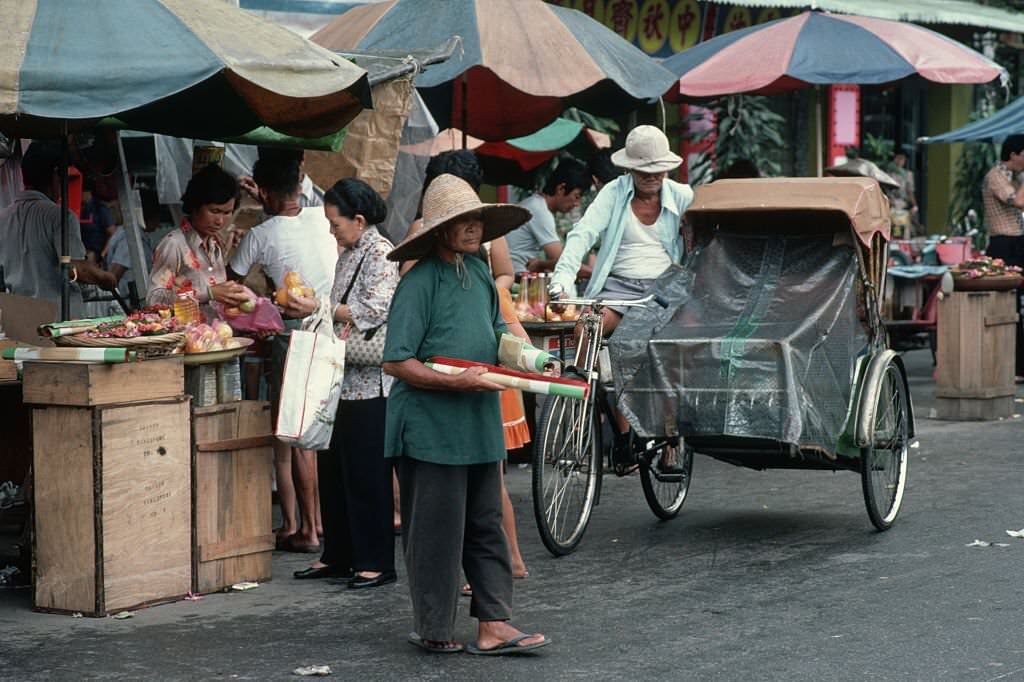 A woman stands next to a man parking a bicycle rickshaw at a market in Singapore.