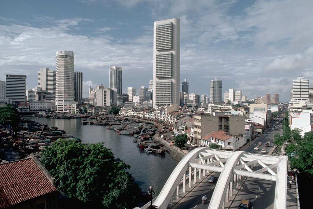 Boats are docked at a quay in the Singapore River near a bridge and a group of skyscrapers, 1980s