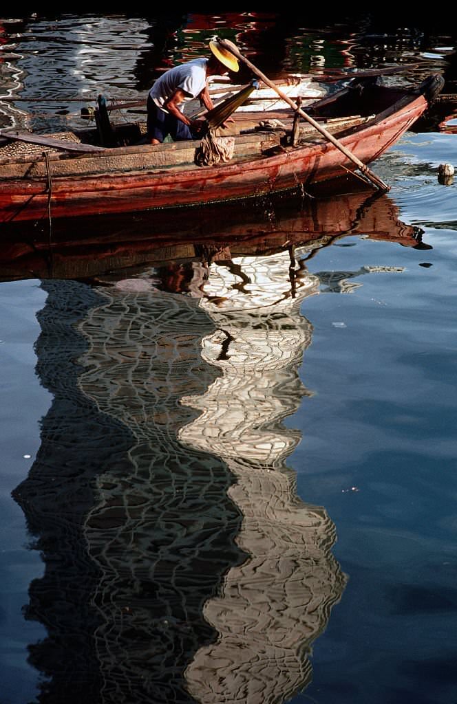 A fisherman prepares his boat by a reflection of a skyscraper in Singapore, 1980s