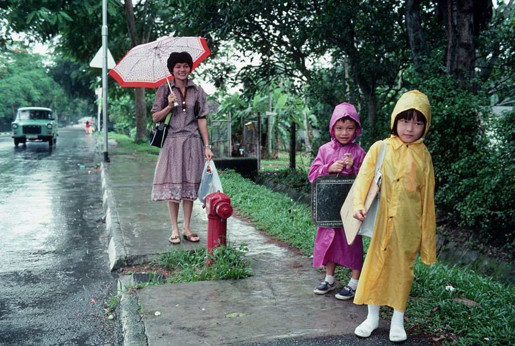 Two school children wait at a bus stop in Singapore, accompanied by a woman who is possibly their mother, 1980s