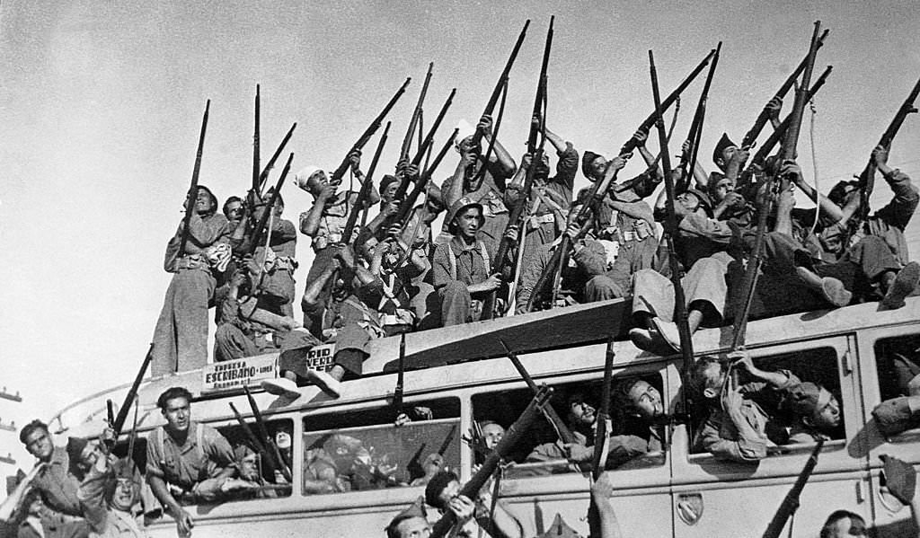 At the Extremadura Front in Spain, a bus full of Republican militiamen waving their arms leaves for the front line. They are for the most part aiming at the sky.