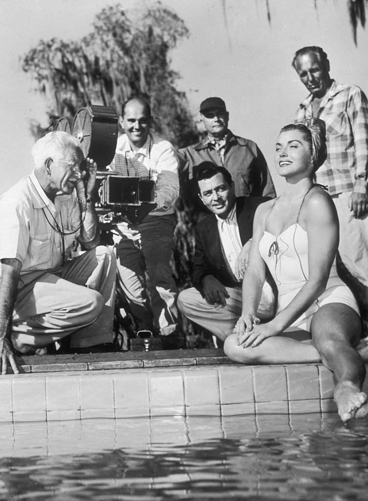 American actor Esther Williams sits in a swimsuit at the edge of a swimming pool, with a camera crew filming her in the background, Cypress Gardens, Florida, 1945