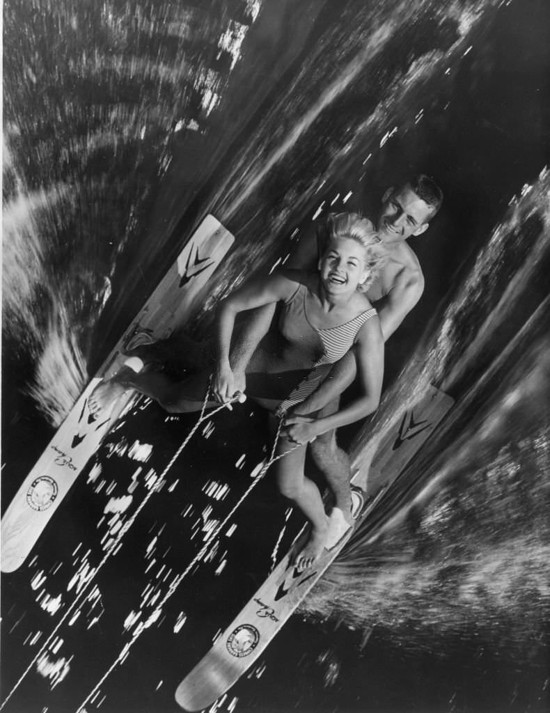 Two water-ski champions on one pair of skis captured by a photographer in an overhanging tree as they speed past at Cypress Gardens, Florida, 1966