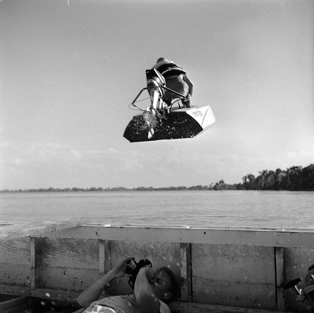 A photographer lies in the bottom of his boat in order to capture an action shot of a speedboat flying overhead, at the Cypress Gardens race course, Florida, 1956