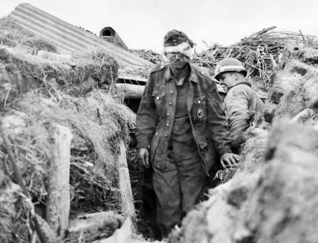 Badly battered German gunner of a gun nest on beach of northern France staggers out of underground nest under watchful eye of a burly U.S. Army M.P. on June 10, 1944.
