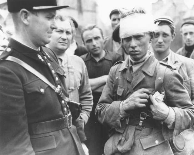 A wounded German soldier who surrendered to the Allied invasion forces, stands surrounded by a crowd of civilians and a French gendarme on the left, in Barneville in the Normandy region, June 1944.
