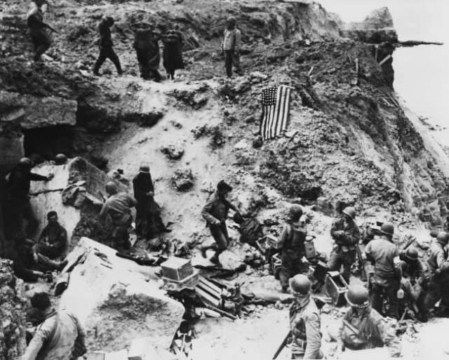 Hands clasped over their heads, a group of German prisoners (top) is herded down a slope on the French coast on June 21, 1944, to a ship to take them to a prison camp, as American command post bustles with activity below.