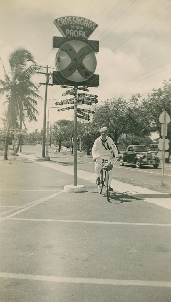 Sailor rides bicycle by Crossroads of the Pacific sign, 1940s