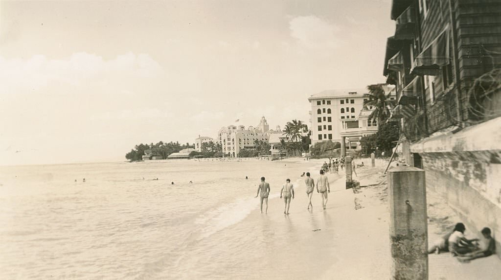 Men in bathing suits walk near surf. Buildings line beach, 1940s