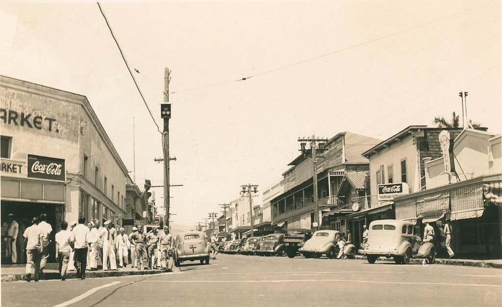 Sailors on a broad commercial street in Hawaii with cars parked on either side, 1940s