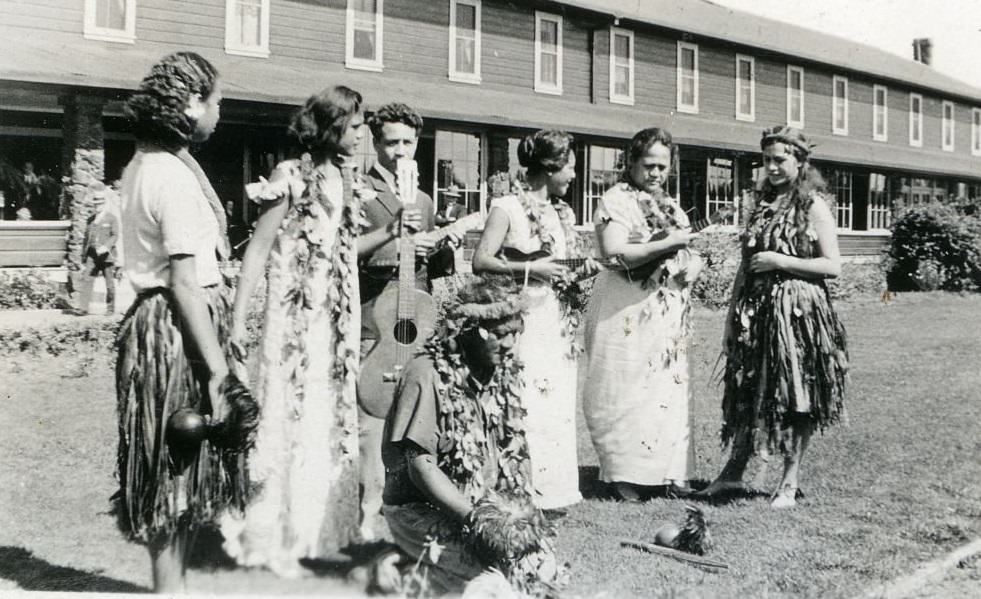 Women wearing leis and hula skirts standing on grass in front of building holding musical instruments, 1940s