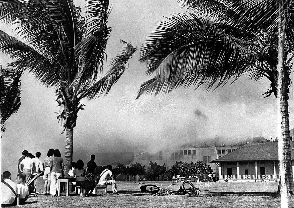 Hawaiians watch as a school building used as an emergency hospital burns fiercely after a heavy bombing attack by Japanese raiders in Honolulu, World War II, 30th December 1941,