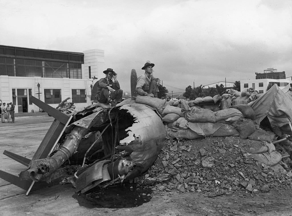 Two servicemen sit on the wreckage of a bomber, surrounded by dirt and sandbags, on Hickam Field after the Japanese attack on Pearl Harbor, Honolulu, Hawaii, 1941