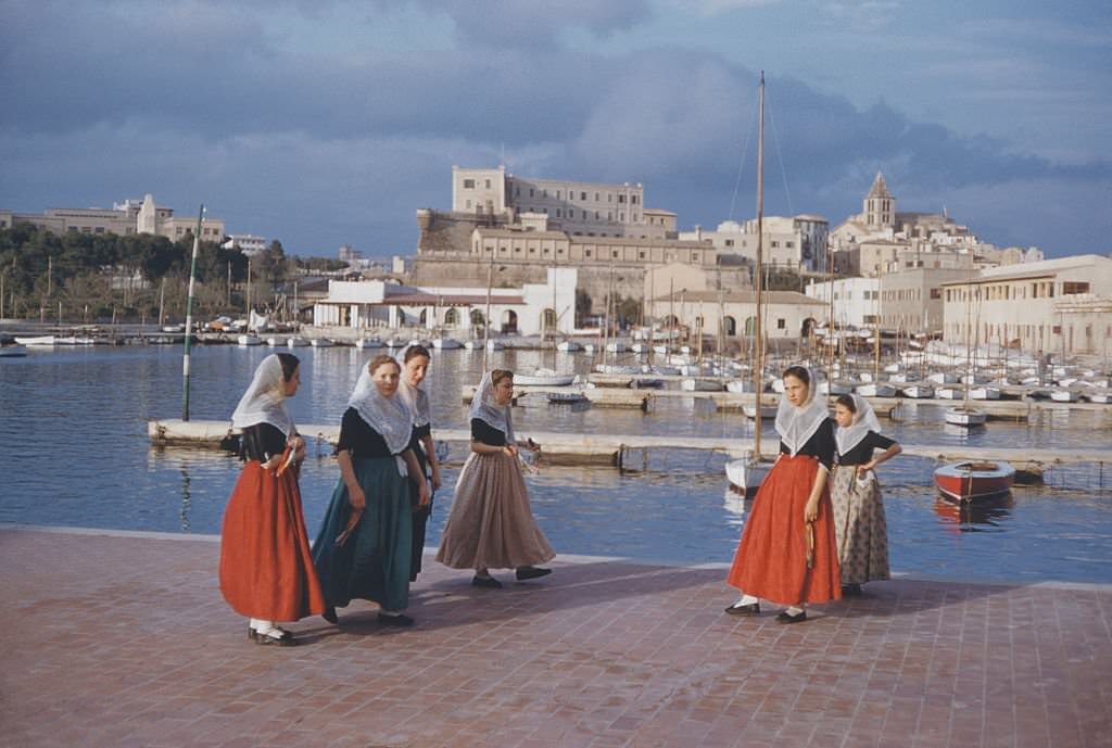 Women and girls in traditional dress walking by the harbour in Majorca, Spain, 1960.