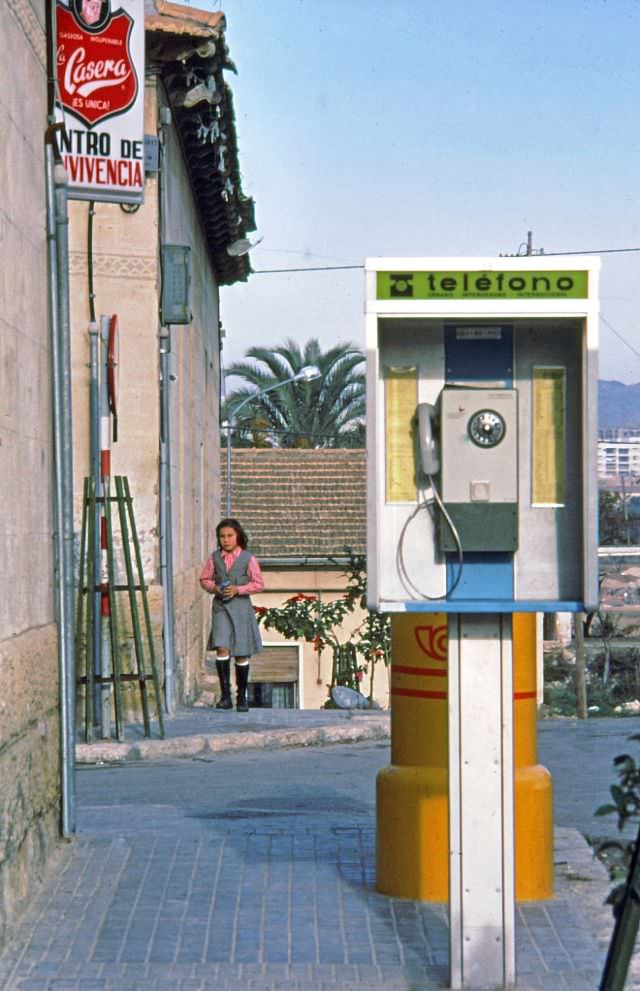 A school girl in her uniform walking towards a mailbox and pay telephone, Barrio Benalúa, Alicante, Valencia, 1977