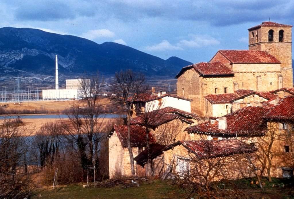 Mid shot of medieval stone buildings and leafless trees, with the Ebro river, Santa Maria de Garona Nuclear Power Plant, and mountains in the background, Burgos, Spain, 1970.