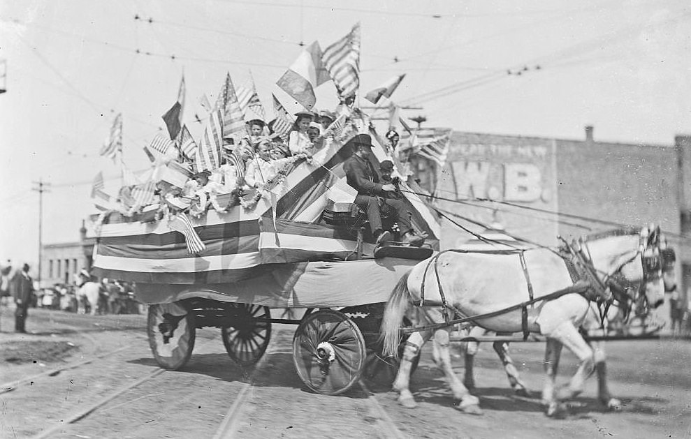 Wagon carrying girls waving flags, moving down a street during a church picnic parade celebrating the feast of St John the Baptist in the Brighton Park community area, Chicago, 1903