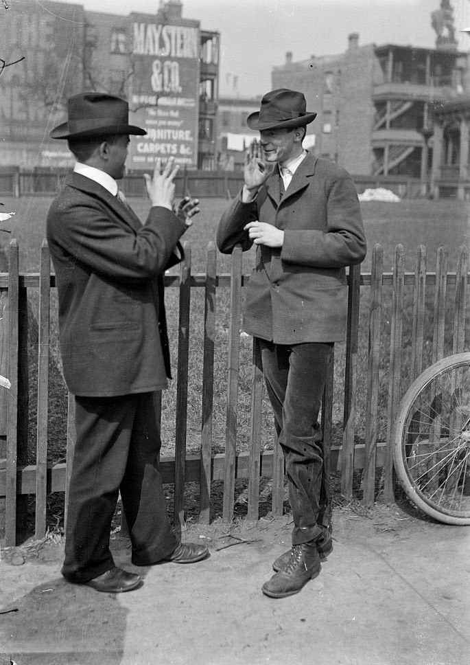 Two deaf men standing in front of a fenced yard or field signing to each other during the Automatic Electric Telephone Company strike, Chicago, Illinois, April 23, 1903.