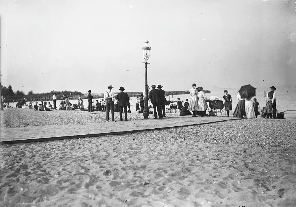 People standing on a boardwalk and in the sand at the 99th Street beach, looking out at Lake Michigan, in the East Side community area of Chicago, Illinois, 1905.