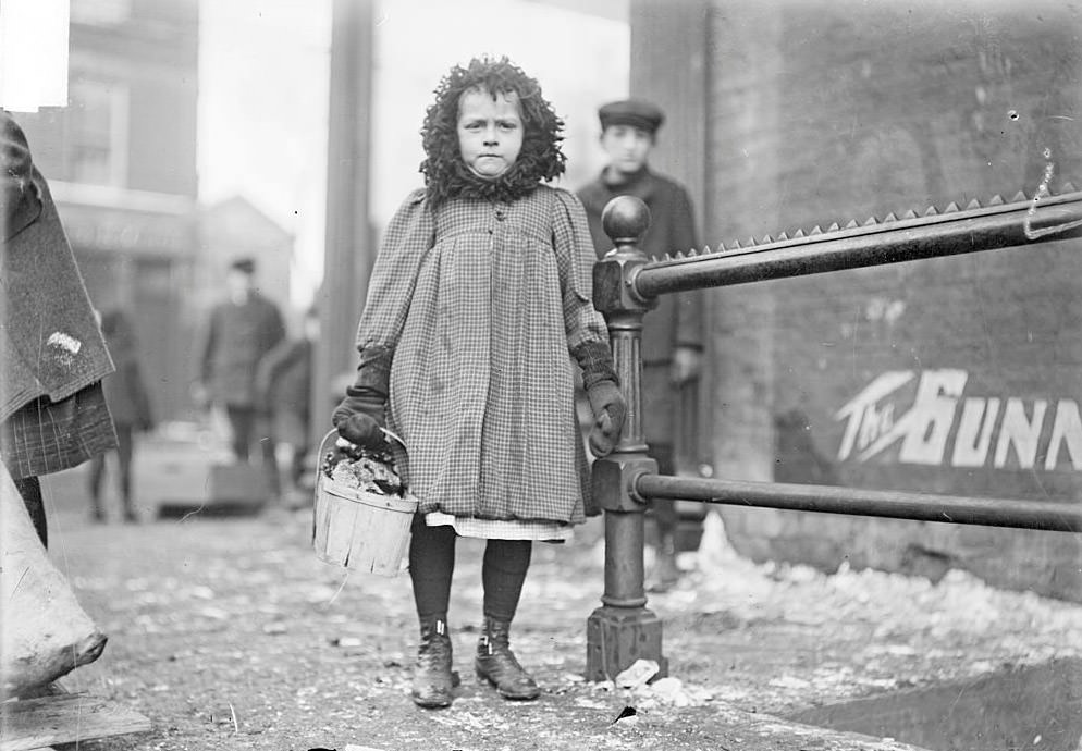 Girl holding a basket of coal on a street corner by the Salvation Army salvage store with other children and pedestrians in the background, Chicago, Illinois, 1903.