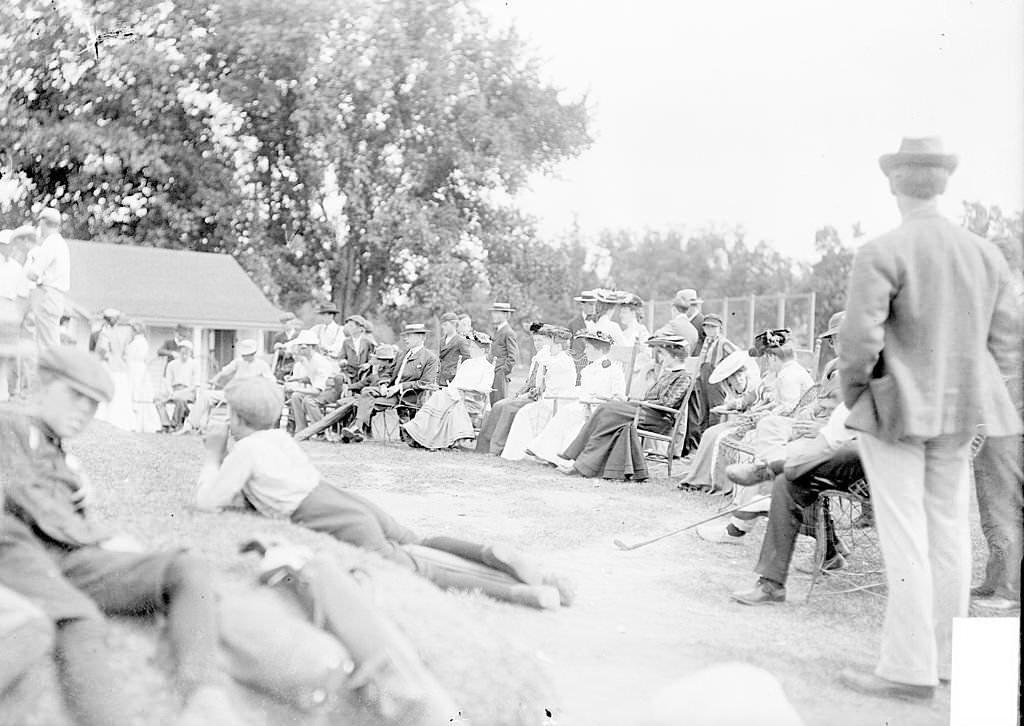 A group of spectators sitting outdoors on the grass in chairs in an open semi-circle, with the men wearing suits, and the women wearing long skirts and hats, at the Exmoor Golf Club located at 700 Vine Avenue in Highland Park, Illinois, 1903.