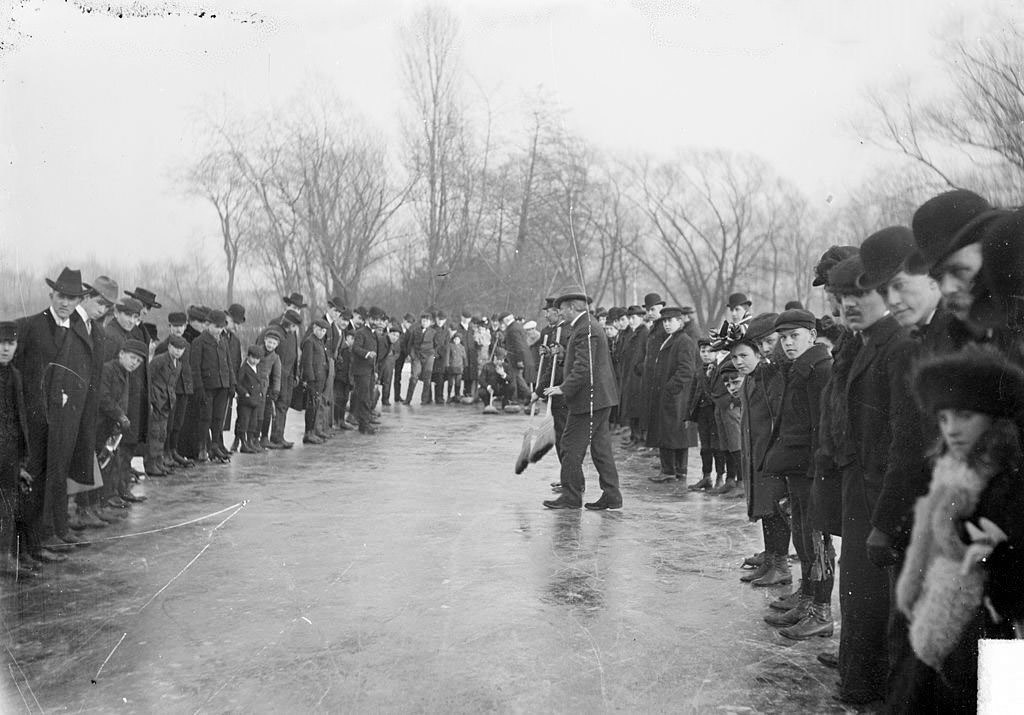 Two men with brooms standing on the ice amidst spectators in a curling match at Jackson Park in Chicago, Illinois, 1903.
