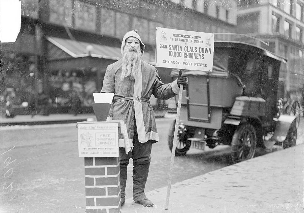 A man dressed up as Santa Claus holding a sign that reads, Help the Volunteers of America to send Santa down 10,000 chimneys of Chicago's poor people, on the sidewalk of a commercial street, Chicago, 1902