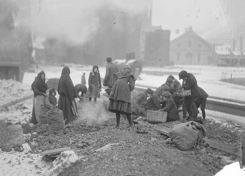 Women and children picking up coal from the ground at the coal yards during winter, Chicago, Illinois, 1902.
