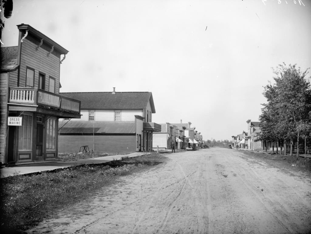 View down dirt road of buildings and storefronts along a sidewalk, possibly Main Street, looking north toward the Chicago & Northwestern railroad tracks, Merrillan, 1901