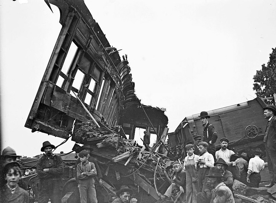 Overturned and damaged train cars from a train accident with men and children standing in front of the wreckage, 1900s.