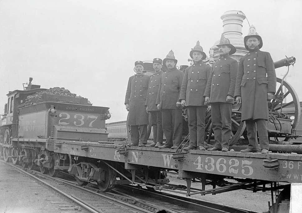 Six firemen standing in front of a fire engine on a train flatcar, 1900s.