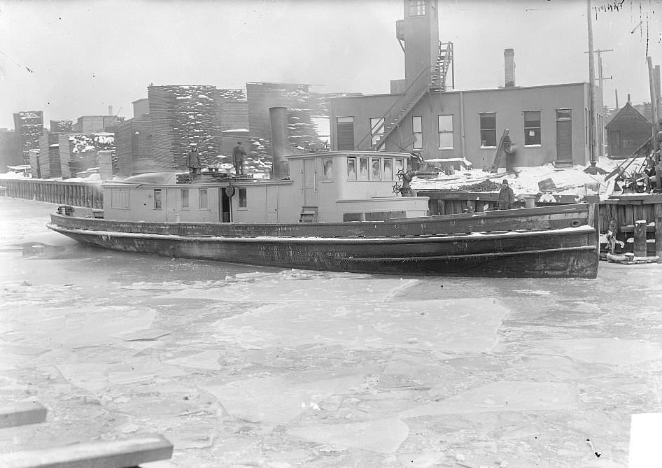 Fireboat, the Swenie, with three men standing on its decks, docked on the Chicago River in front of an industrial building with boards stacked nearby, Chicago, Illinois, 1900s.