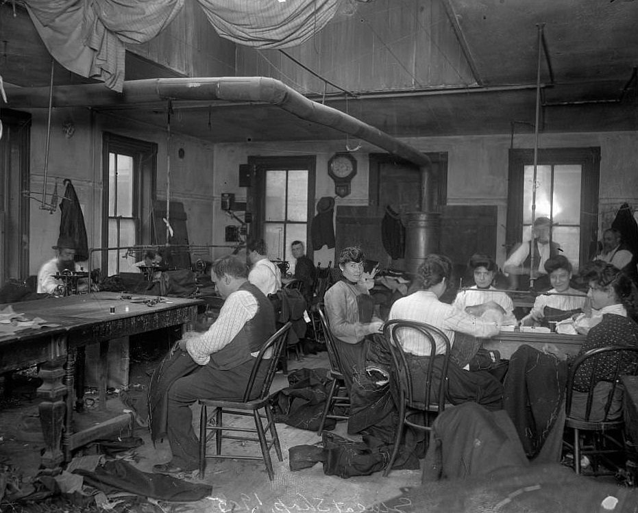 Women and men sewing at various tables and sewing machines in the workroom of a sweatshop at 132 Maxwell Street in the Near West Side community area, Chicago, Illinois, 1905.