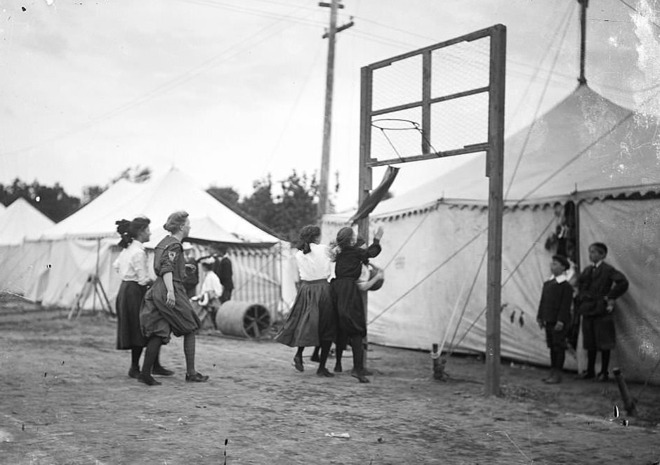 Girls playing basketball on a grass court surrounded by light colored tents, Chicago, Illinois, 1905.