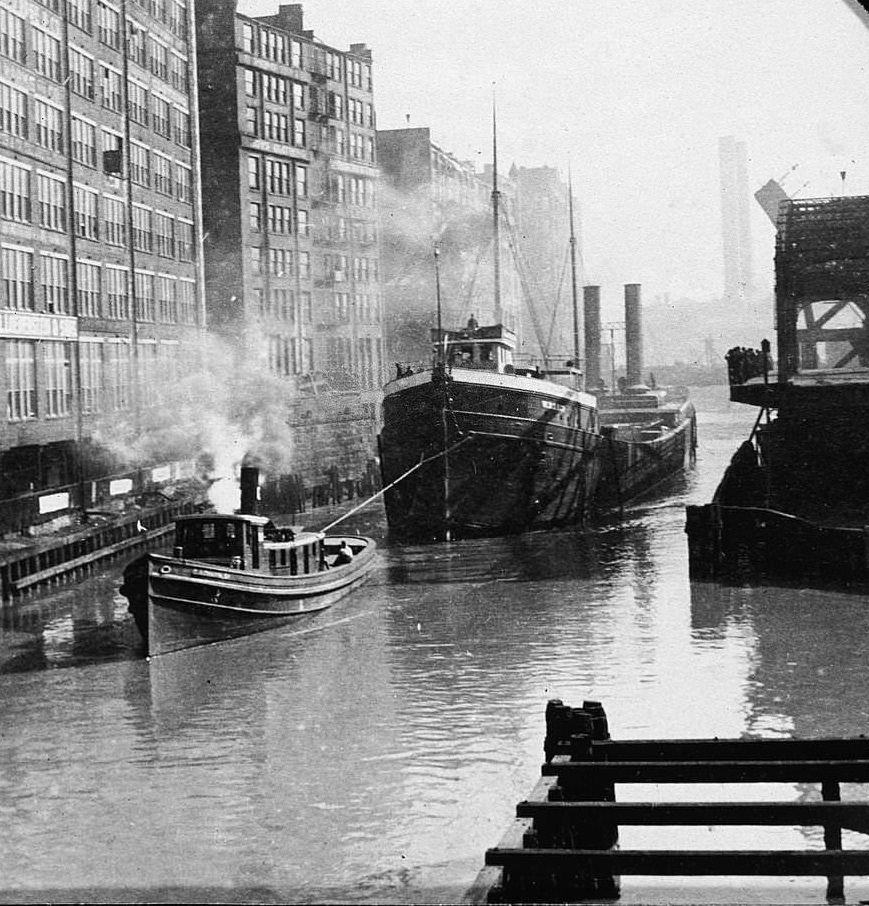 A tugboat at work pulls a larger boat along the Chicago River waterfront, Illinois, 1905