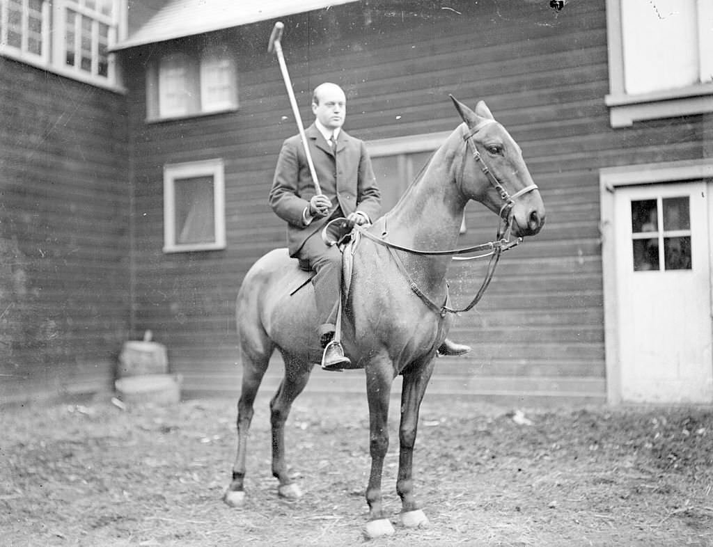 Polo player holding a polo mallet, sitting on horseback in a paddock area in front of a barn in Chicago, Illinois, 1901.