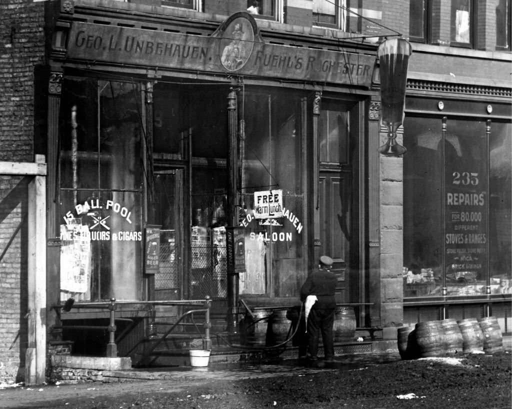 A man using a hose to wash the windows of the George L. Unbehauen Saloon, next to the Northwestern Stove Repair Company Building, Chicago, IL, 1900.