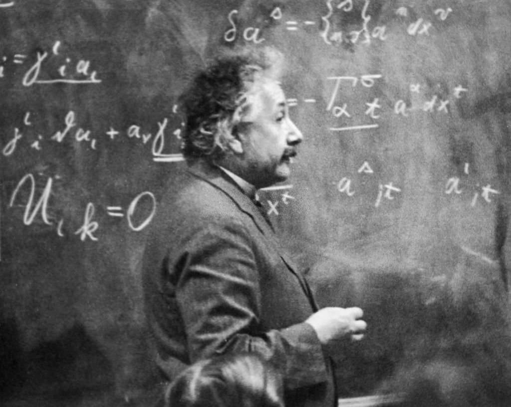Albert Einstein standing beside a blackboard with chalk-marked mathematical calculations written across it, 1931