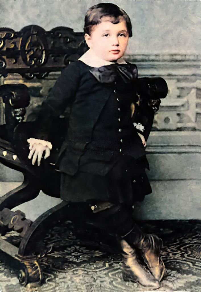 Three-year-old Albert Einstein, at approximately three years old, wearing a black suit and large neck bow, leaning against an X-chair, and facing the camera with a calm expression, 1882.