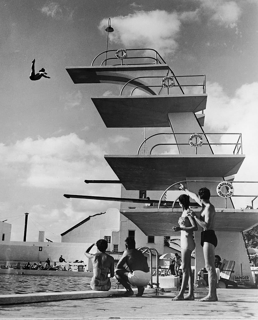 People sit poolside and watch as a swimmer dives off the high platform of the Olympic-size swimming pool at the MacFadden - Deauville Hotel, 1950s