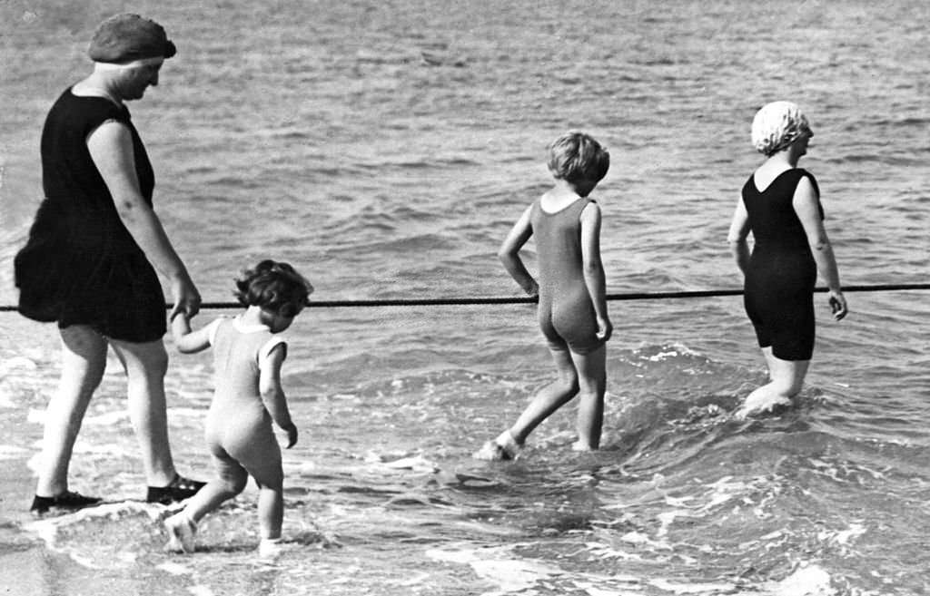 A woman and her children wearing one-piece swimming-suit secure themselves holding a rope as they enter in the water, 1937