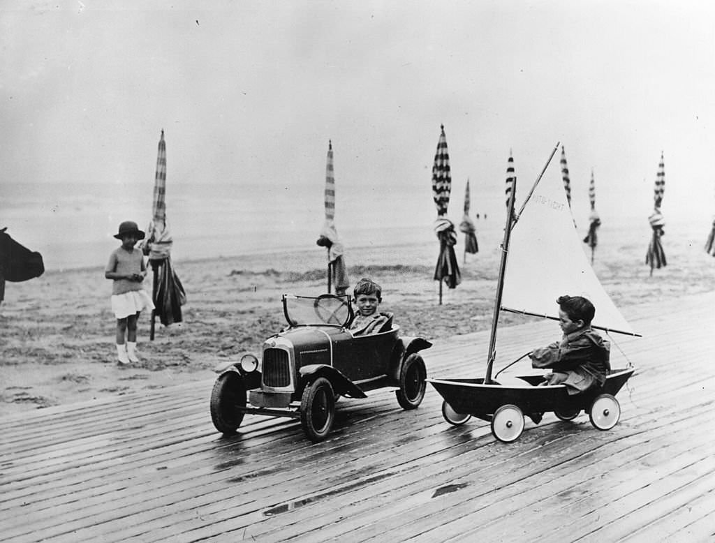 Two children racing each other in a toy car and boat, along the beach front in Deauville, 1920s