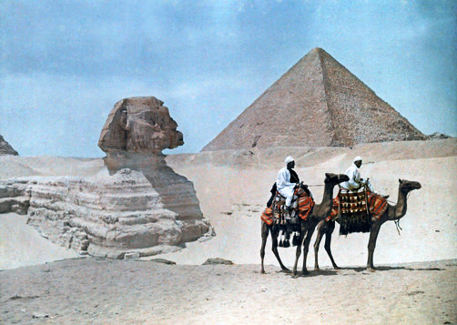 Two men on camel-back ride past the Sphinx and Great Pyramid.