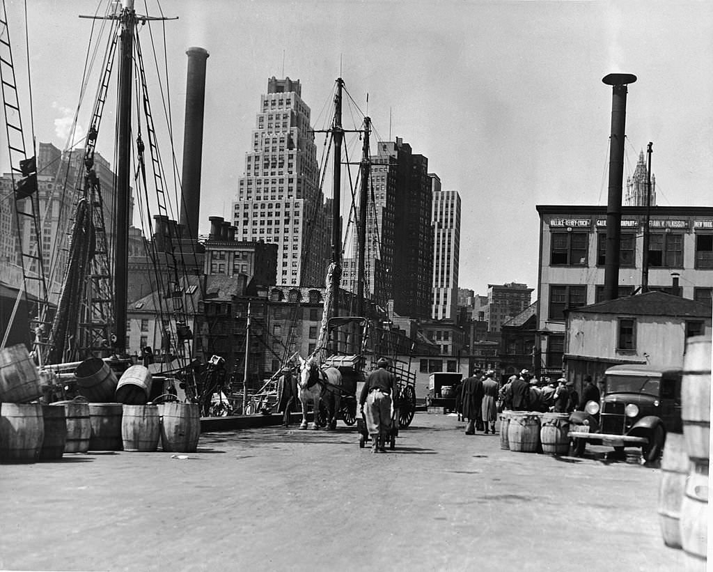 View of the East River docks, New York, New York, 1930s. The masts of several docked fishing boats are visible at left.