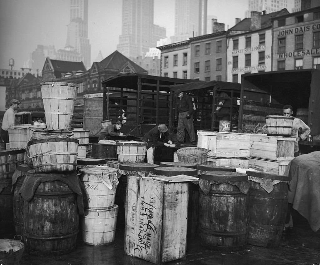 A view of men working behind stacks of wooden crates and barrels at the Fulton Street Fish Market in downtown Manhattan, New York City, 1920s