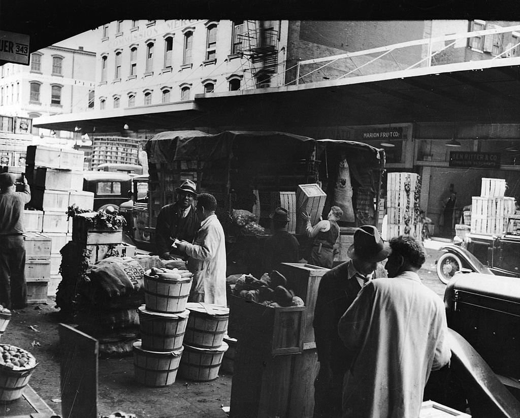 en congregate around bushels and sacks of fruits and vegetables at the Fulton Street Market as a workers stack and unload crates from a truck, 1930s.