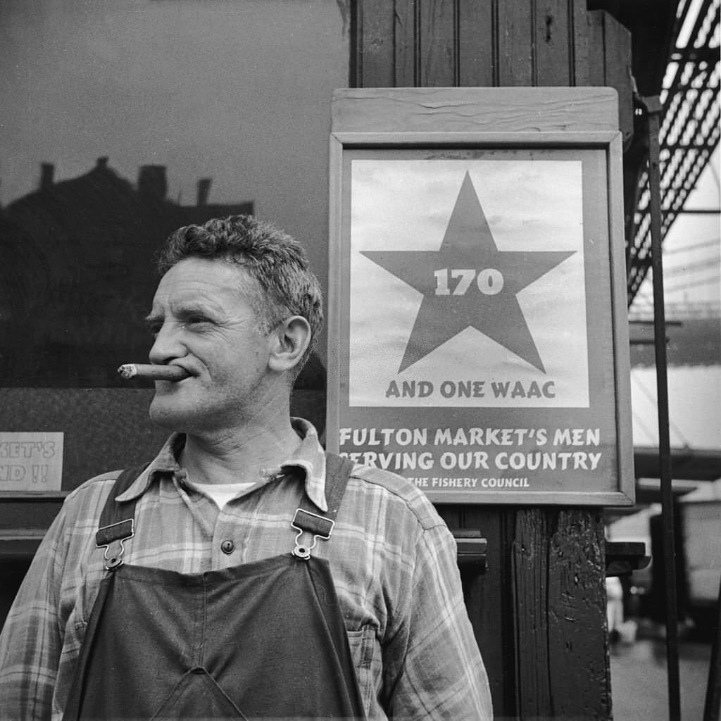 Man smokes a cigar while standing next to a World War II sign at the Fulton fish market, New York City, 1943