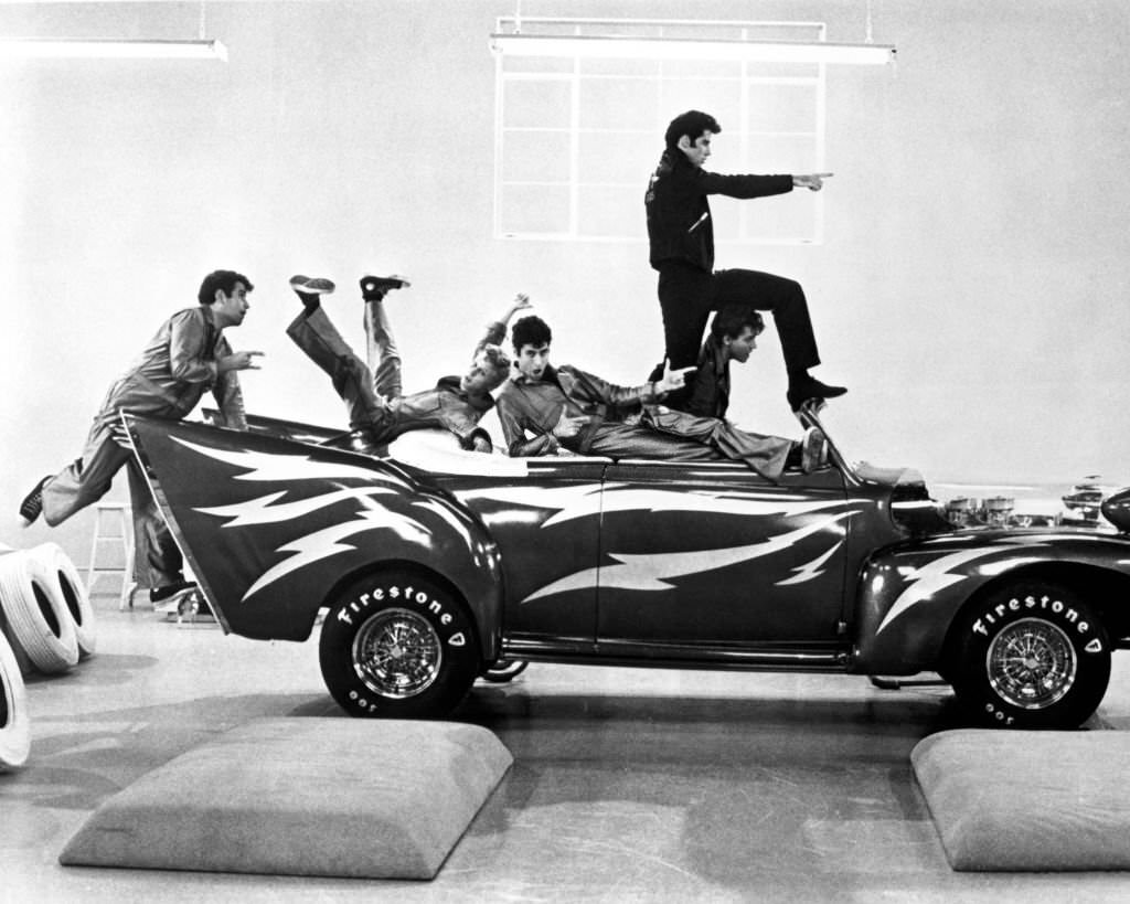 John Travolta as Danny, posing on a 1948 Ford DeLuxe convertible with the rest of the T-Birds in the 'Greased Lightnin'' scene from the musical film 'Grease', 1978.