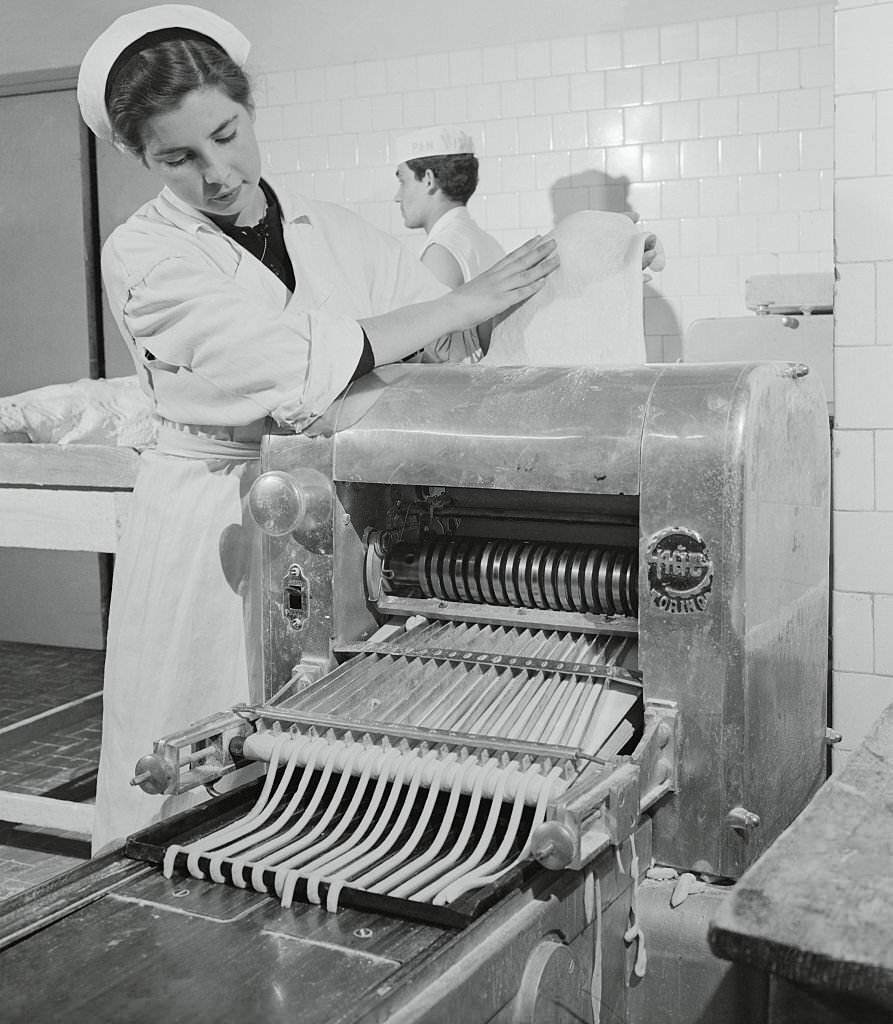 Woman Feeding Dough into a Machine. Passing through mechanical channels, the future bread sticks resemble thick spaghetti as they begin to take on an elongated appearance.